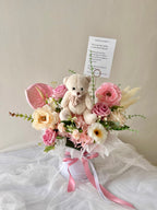 Bloombox with pink flowers, a teddy bear, and a card on a white surface.