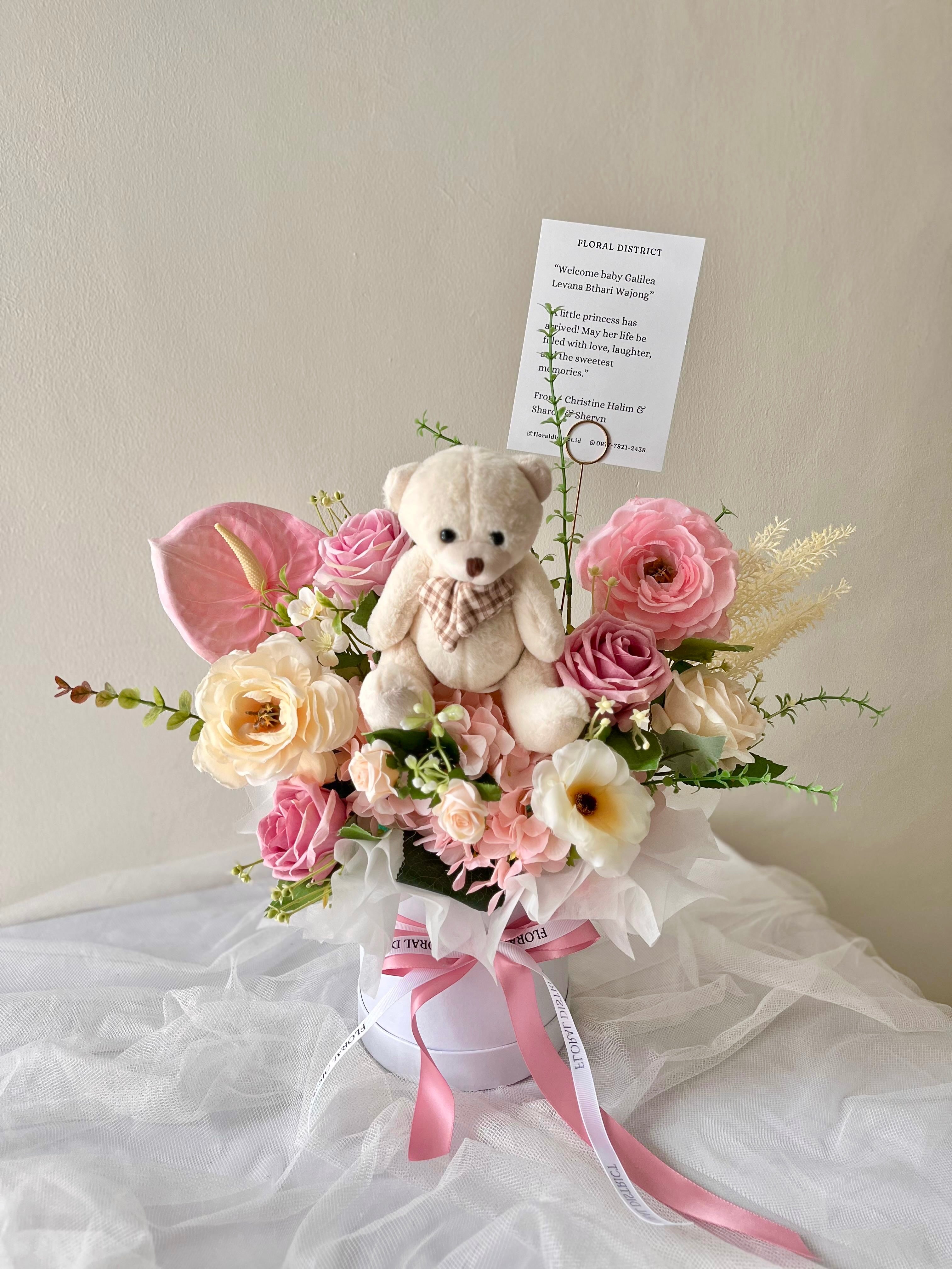 Bloombox with pink flowers, a teddy bear, and a card on a white surface.