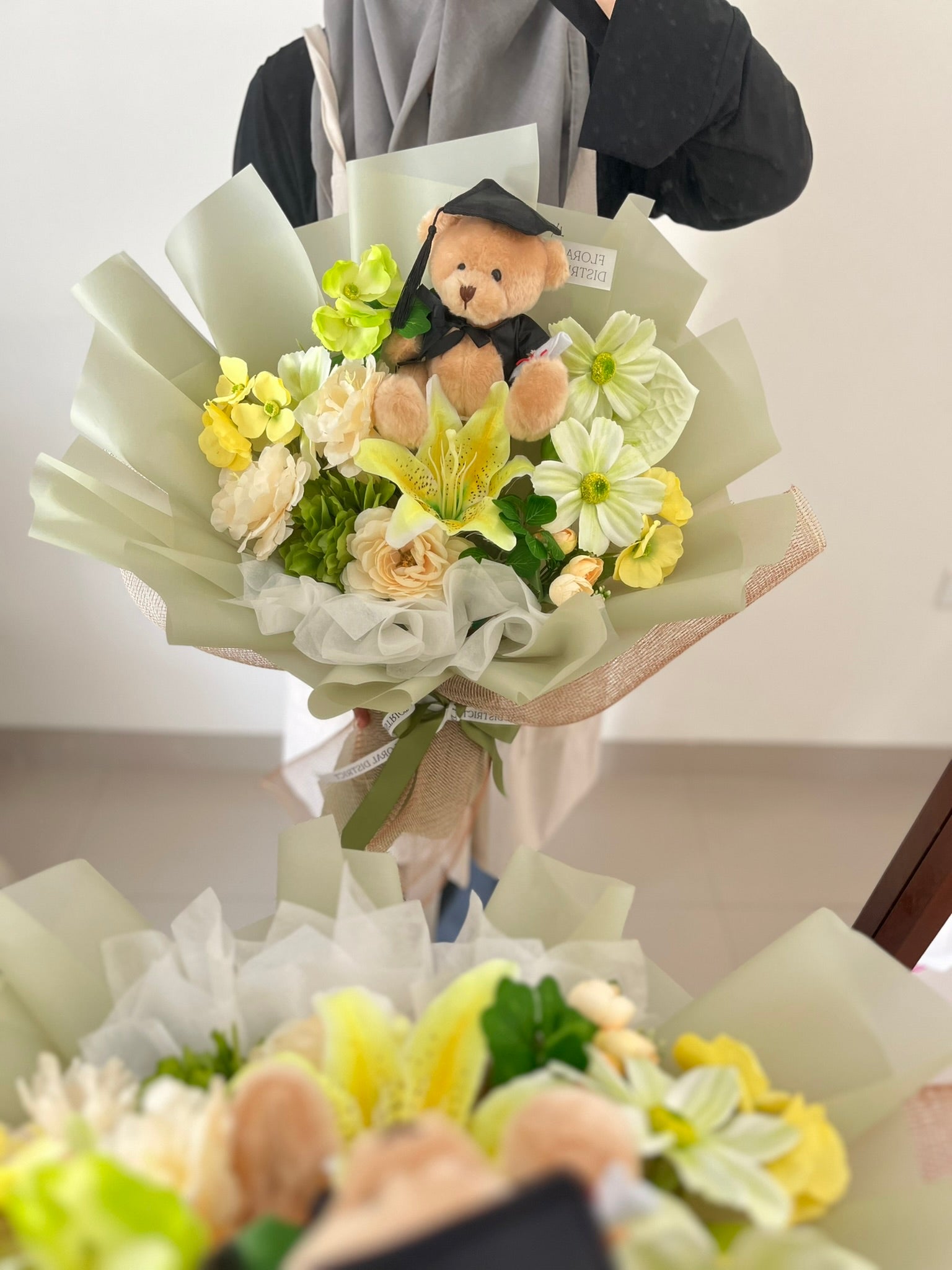 Bouquet of flowers with a teddy bear in a graduation cap and gown, held by a person.